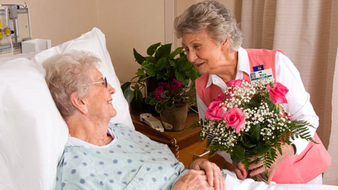 An elderly volunteer delivering flowers to an elderly patient in a hospital room.