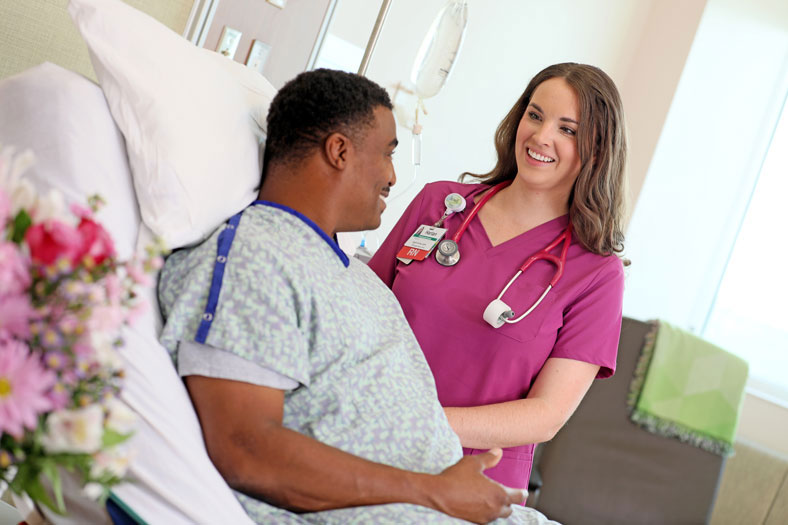 Nurse in pink scrubs greeting a patient in bed with a friendly smile.