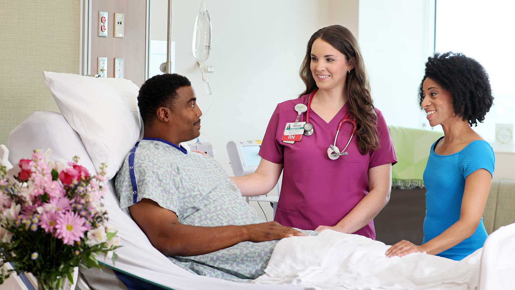 A nurse checks on a patient as his wife sits by his side.