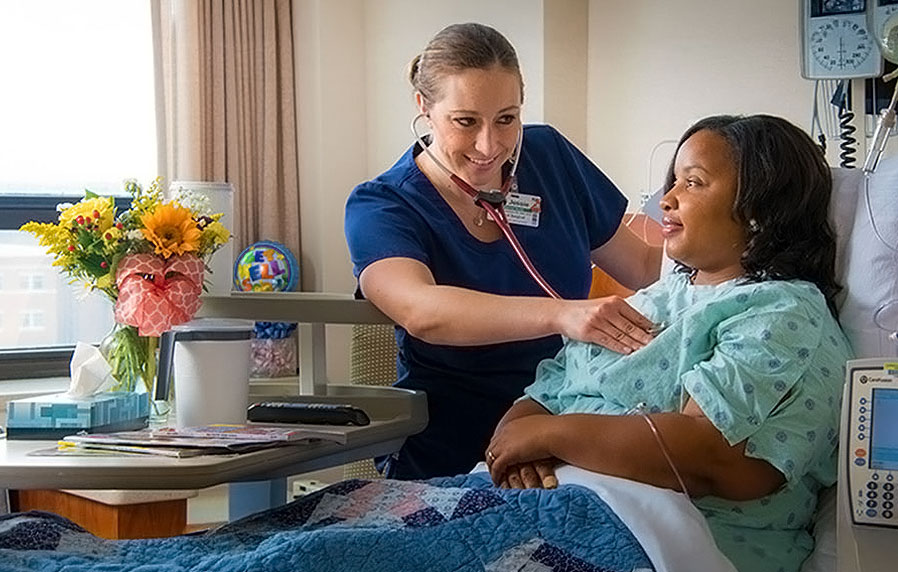 A smiling nurse listening to a woman's heartbeat in a hospital room.