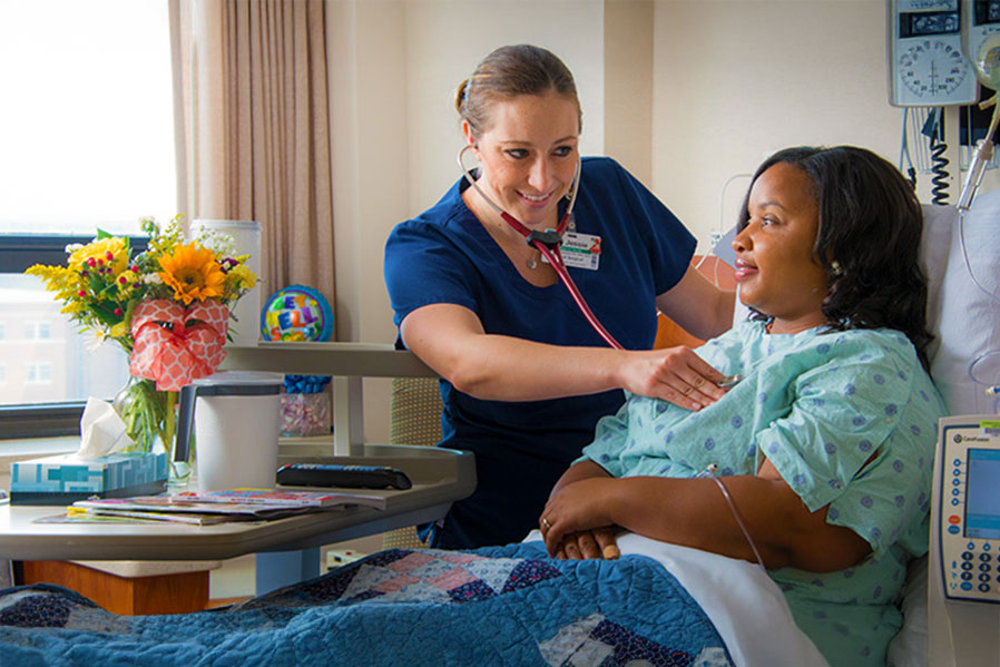 A smiling nurse listening to a woman's heartbeat in a hospital room.